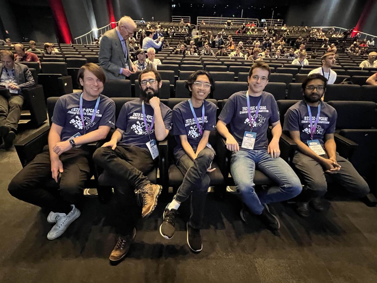 5 young men in jeans and tshirts sitting in front row of auditorium.