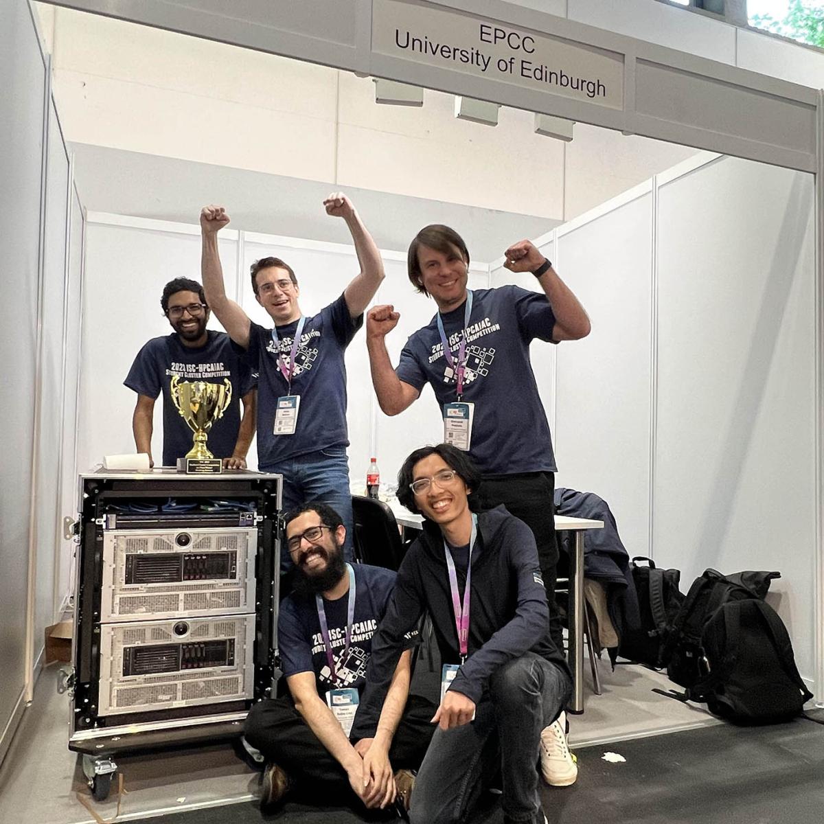 5 triumphant young men in jeans and t-shirts standing beside computer cluster with gold trophy cup on top